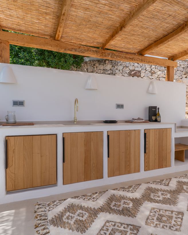 Buitenkeuken met houten fronten onder een rieten pergola tegen een natuurstenen wand